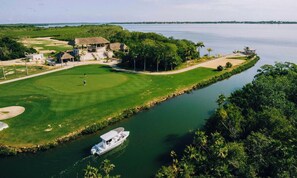 A golf course with a water body, a boat, and a building with a thatched roof. - Placencia Lagoon: 2BR Waterfront Rental (Placencia)