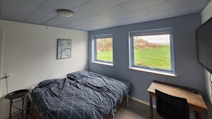 Ein Schlafzimmer mit Bett, Schreibtisch und zwei Fenstern mit Blick auf ein Wiesenfeld.