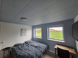 Un dormitorio con cama, escritorio y dos ventanas que dan a un campo cubierto de hierba.