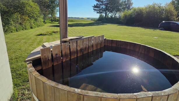Ein hölzernes Whirlpool mit Metallstange, umgeben von einer Rasenfläche mit Bäumen und blauem Himmel.