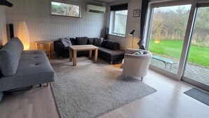 Ein modernes Wohnzimmer mit einem Ecksofa, einem kleinen Tisch und einem Sessel. Ein großes Fenster bietet Blick ins Freie.