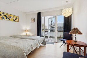 Ein Hotelzimmer mit Bett, Sessel, Tisch, Lampe, einem Gemälde und Blick durch eine Glastür.