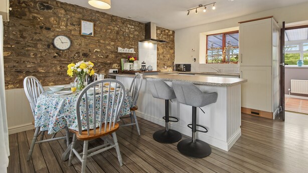 Una cocina con pared de piedra, suelo de madera, una mesa de comedor con sillas, una barra y vistas a través de una ventana.