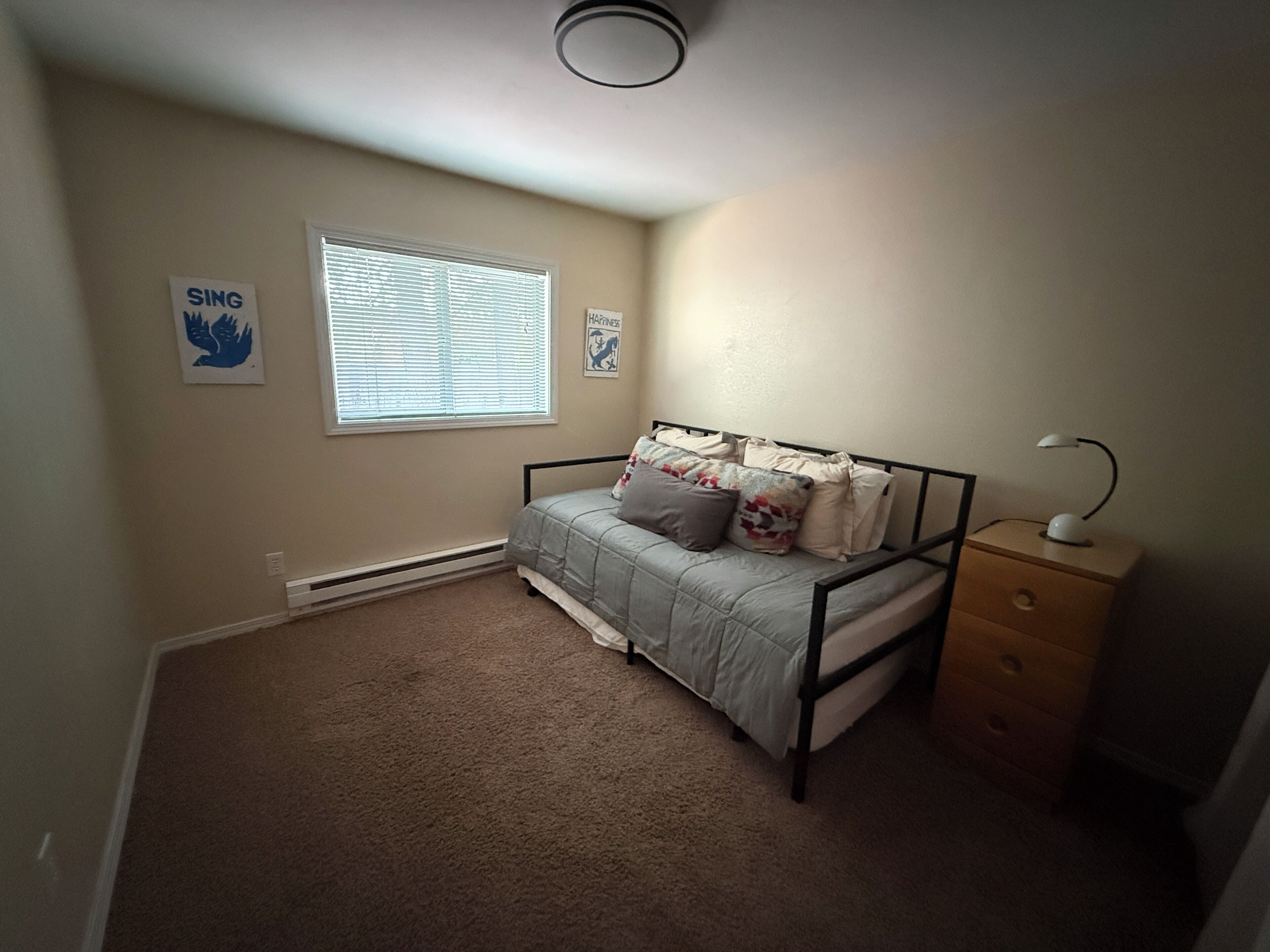 Un dormitorio con cama, mesita de noche, cómoda, ventana con persianas y dos adornos en la pared.