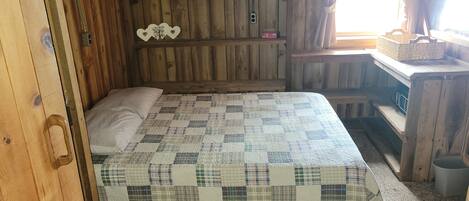 Una habitación de cabaña de madera con una cama, una estantería con adornos y una ventana con cortinas.