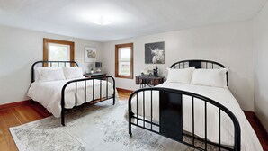 Een slaapkamer met twee zwarte metalen bedden, wit beddengoed, een kamerkleed met patroon en ingelijste kunstwerken aan de muur.