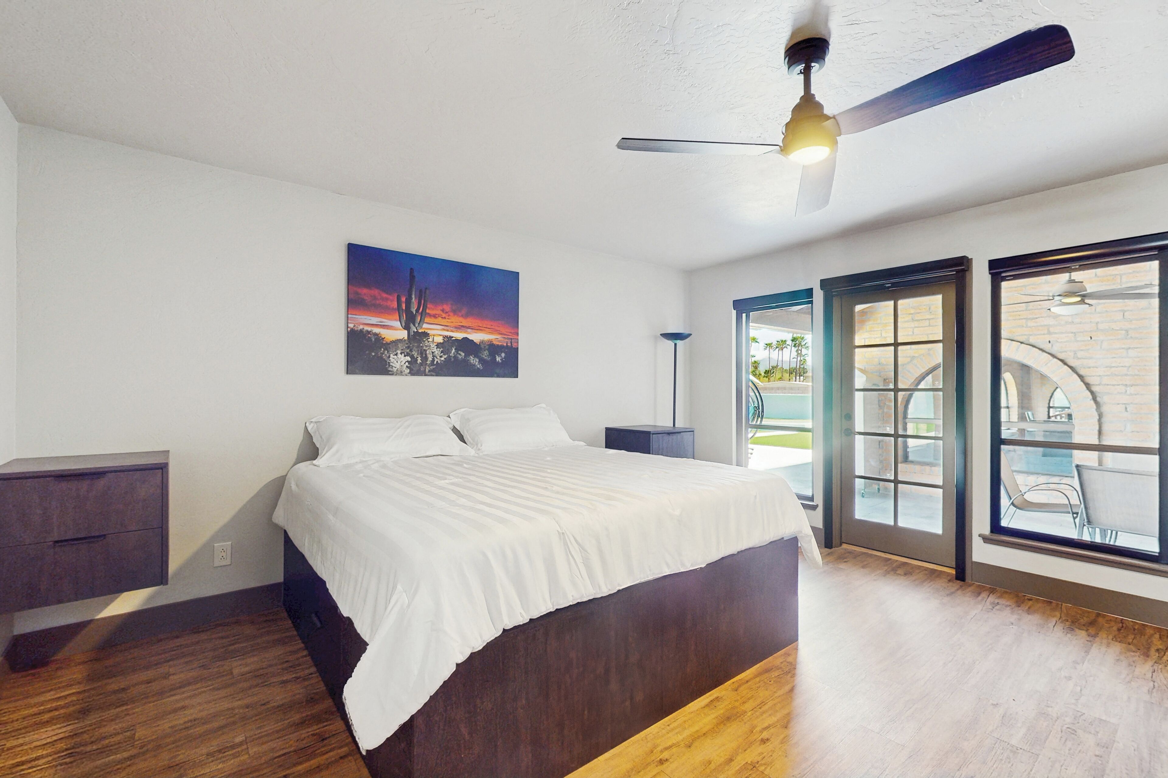 A bedroom with a bed, a nightstand, a ceiling fan, a window with a view, and a door leading to an outdoor area.