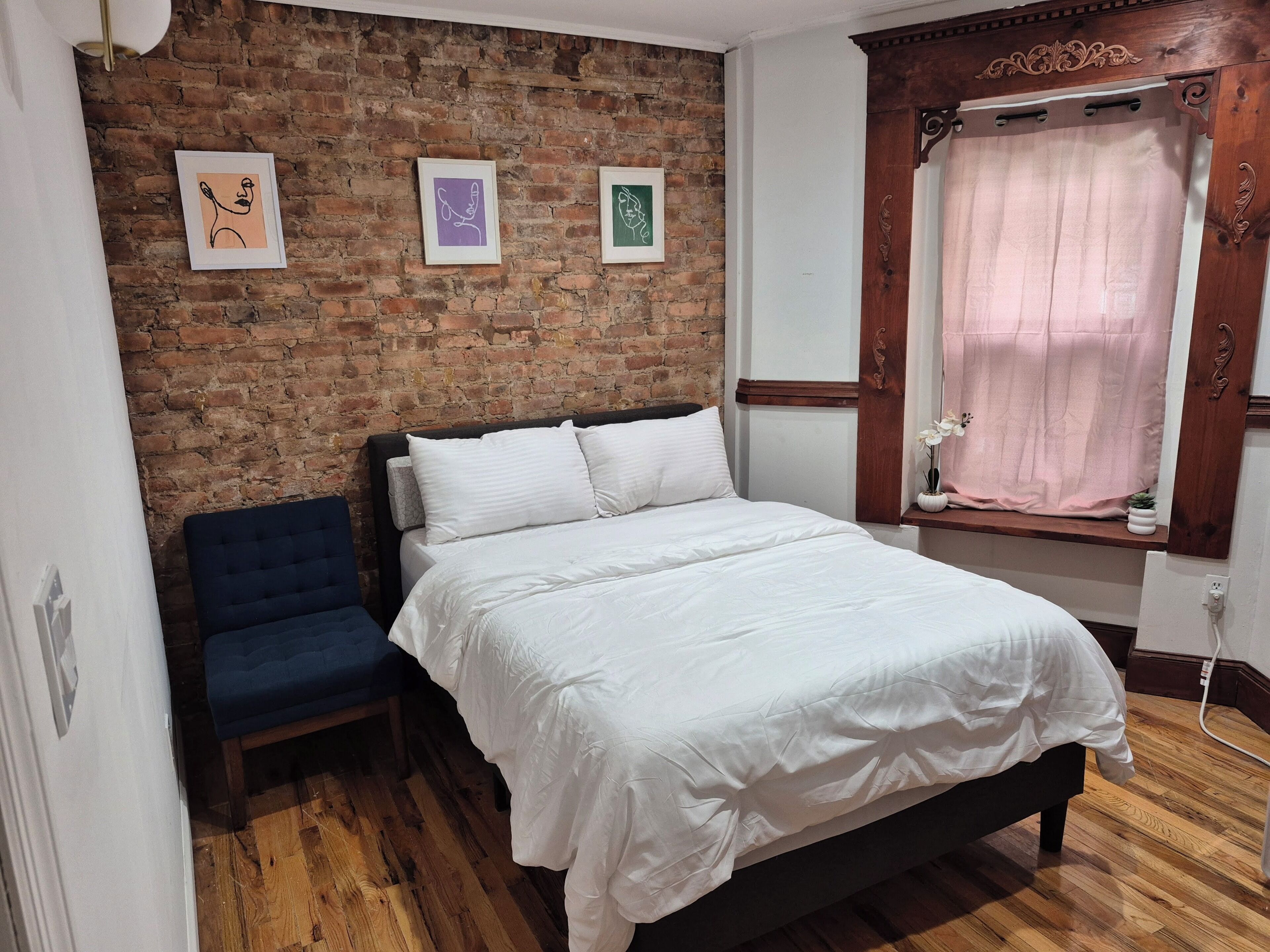 A bedroom with a brick wall, a bed with white bedding, a blue chair, and a window with a curtain.