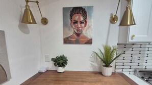 Een kleine keuken met een houten aanrecht, twee blauwe stoelen, een plant in pot en een schilderij aan de muur.