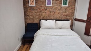 Een slaapkamer met een bakstenen muur, een bed met wit beddengoed, een donkerblauwe stoel en drie ingelijste kunstwerken.