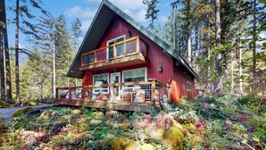 Una cabaña con terraza de madera, amplios ventanales y fachada roja, rodeada de un bosque.