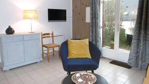A living room with a TV, a lamp, a chair, a sofa, and a window with curtains. - Maisonnette 4 personnes proche de la plage - WIFI (SARZEAU)