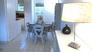 A dining area with a table and chairs, a lamp, and a view of a garden through a window. - Maisonnette 4 personnes proche de la plage - WIFI (SARZEAU)