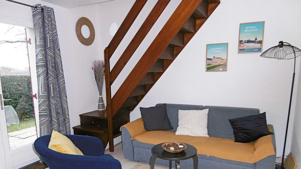 A living room with a sofa, a chair, a small table, a staircase, and a door leading outside. - Maisonnette 4 personnes proche de la plage - WIFI (SARZEAU)