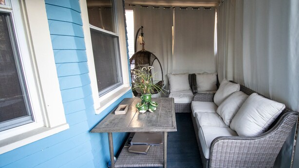 Eine überdachte Veranda mit einem Korbsessel, einem kleinen Tisch mit einer Vase und einem Fenster mit Jalousien.