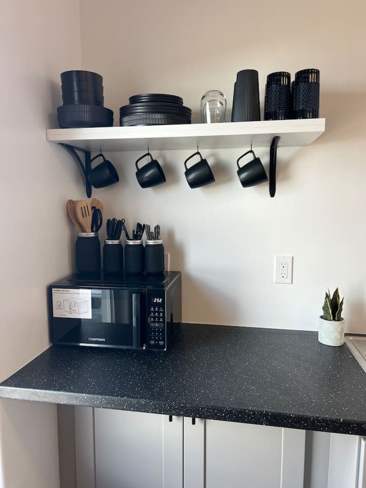 A kitchen with a black countertop, a microwave, a shelf with black kitchenware, and a potted plant.