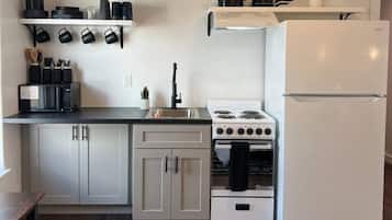 Een kleine keuken met witte kasten, een witte koelkast, een fornuis en een magnetron. Er is een zwart aanrecht en een kamerkleed op de vloer.