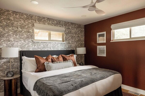 Une chambre à coucher avec un grand lit, deux tables de chevet, un ventilateur de plafond et une fenêtre donnant sur un beau paysage.
