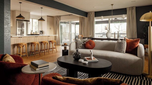 Una sala de estar moderna con un sofá, una mesa de centro y taburetes de bar. Se ve una cocina al fondo con un refrigerador y una ventana.