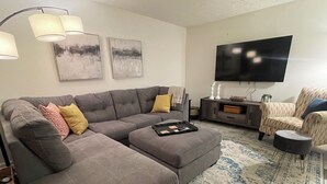 Ein Wohnzimmer mit einem grauen Ecksofa, einem kleinen Hocker, einem gemusterten Teppich, einem Fernseher und einem Beistelltisch mit Dekoration.