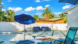 Ein Swimmingpool mit Liegestühlen und einem Sonnenschirm, umgeben von Palmen und einem weißen Zaun.