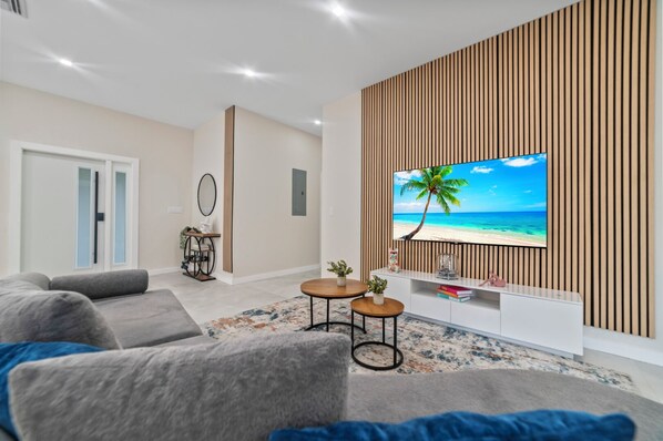 A modern living room with a flat-screen TV on a wooden panel wall, grey sofas, a coffee table, and a small side table with a plant.