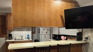 Een kleine keuken met houten kastjes, een wit aanrecht, twee barkrukken, een koelkast, een magnetron en een flatscreen televisie aan de wand.