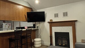 Een ruimtelijk ingerichte leefruimte met een kitchenette, een flatscreen televisie boven de toonbank, een open haard en een zithoek met een bank en stoelen.