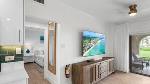 Un salón moderno con un televisor de pantalla plana grande, armarios de madera, un sofá blanco y vistas a una playa a través de una puerta corrediza de vidrio.
