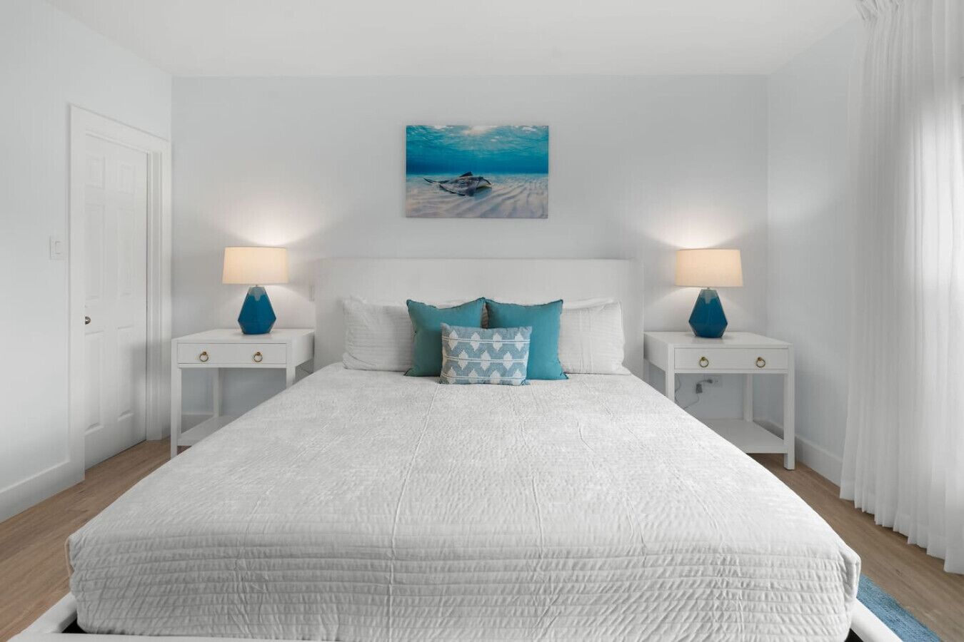 Un dormitorio amplio con una cama grande, dos mesitas de noche con lámparas, un cuadro con una playa y paredes blancas.