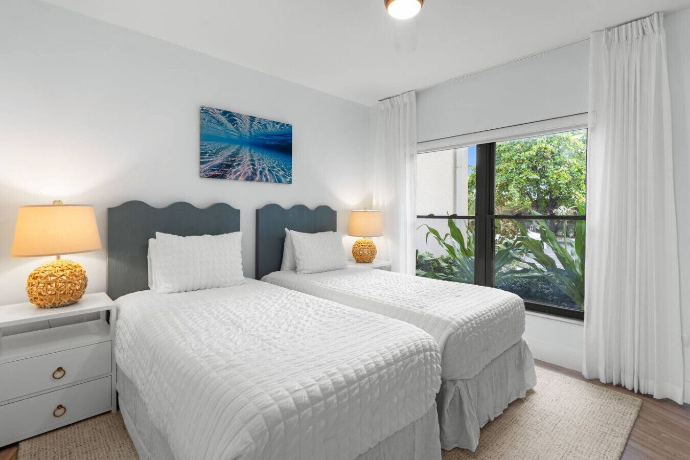A hotel room with two beds, a large window with curtains, a wall-mounted painting, and bedside lamps.