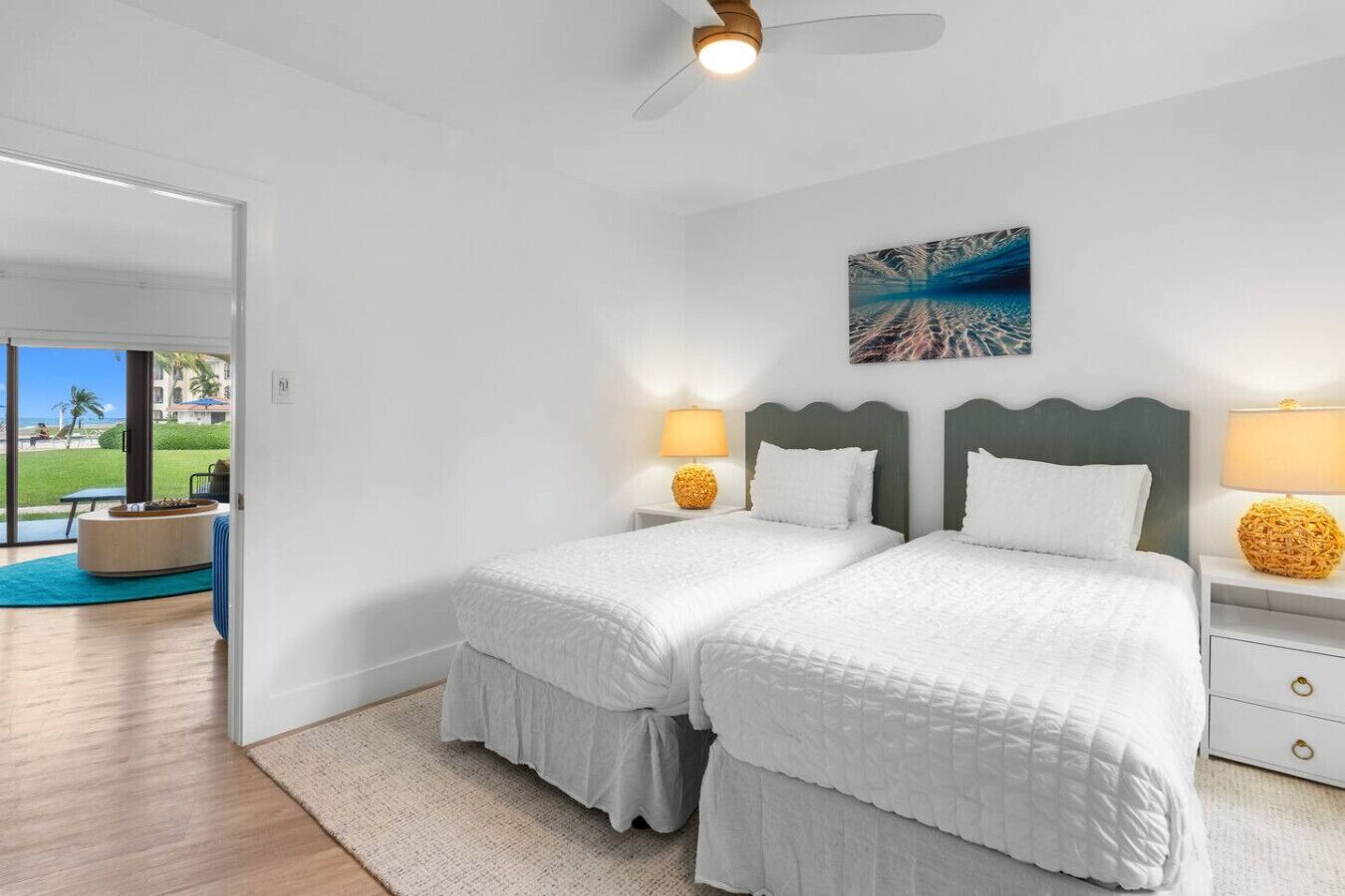 A hotel room with two beds, a ceiling fan, a painting, and a view of the beach.