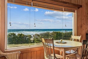 A wooden interior with a large window overlooking the ocean, a round table with chairs, and hanging light fixtures. - Windwhistle - Oceanfront! (Cannon Beach)