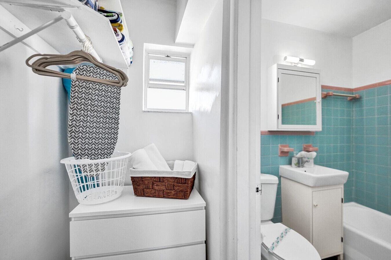 A bathroom with a white toilet, a white sink, and a mirror above the sink. There is a basket on the floor and a hanging rack with towels.
