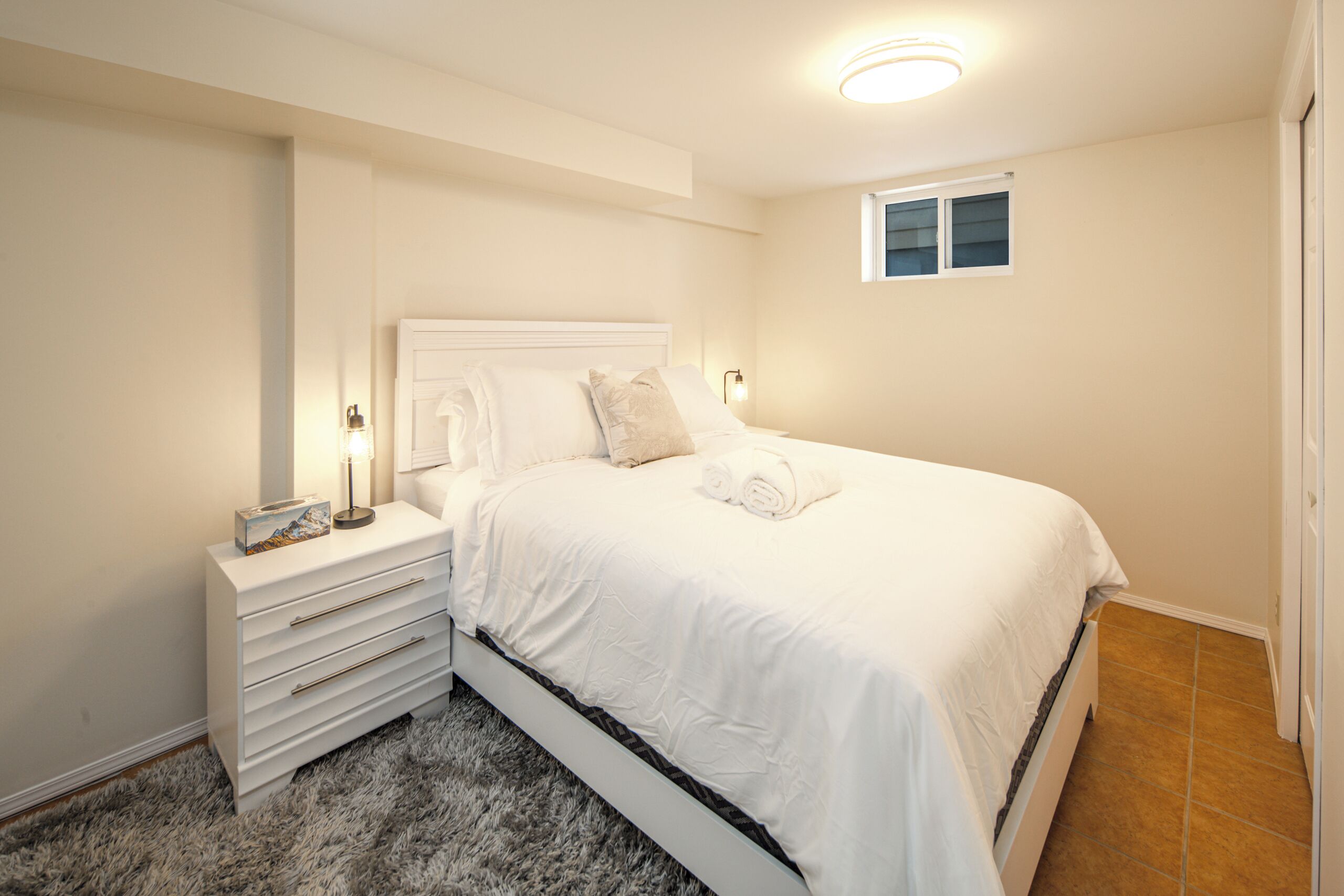 Une chambre à coucher avec un lit blanc, une table de chevet, une lampe et une fenêtre.