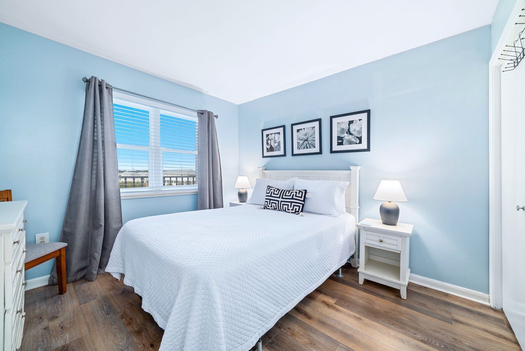 A bedroom with a bed, two bedside tables with lamps, a window with curtains, and three framed pictures on the wall.
