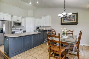 Uma cozinha com armários brancos, uma ilha central, uma mesa de jantar com cadeiras e um lustre.