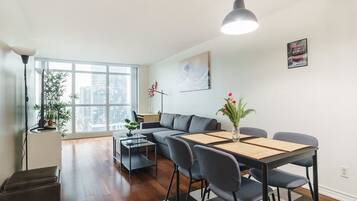 A modern living room with a sofa, dining table, chairs, and a large window offering a city view.
