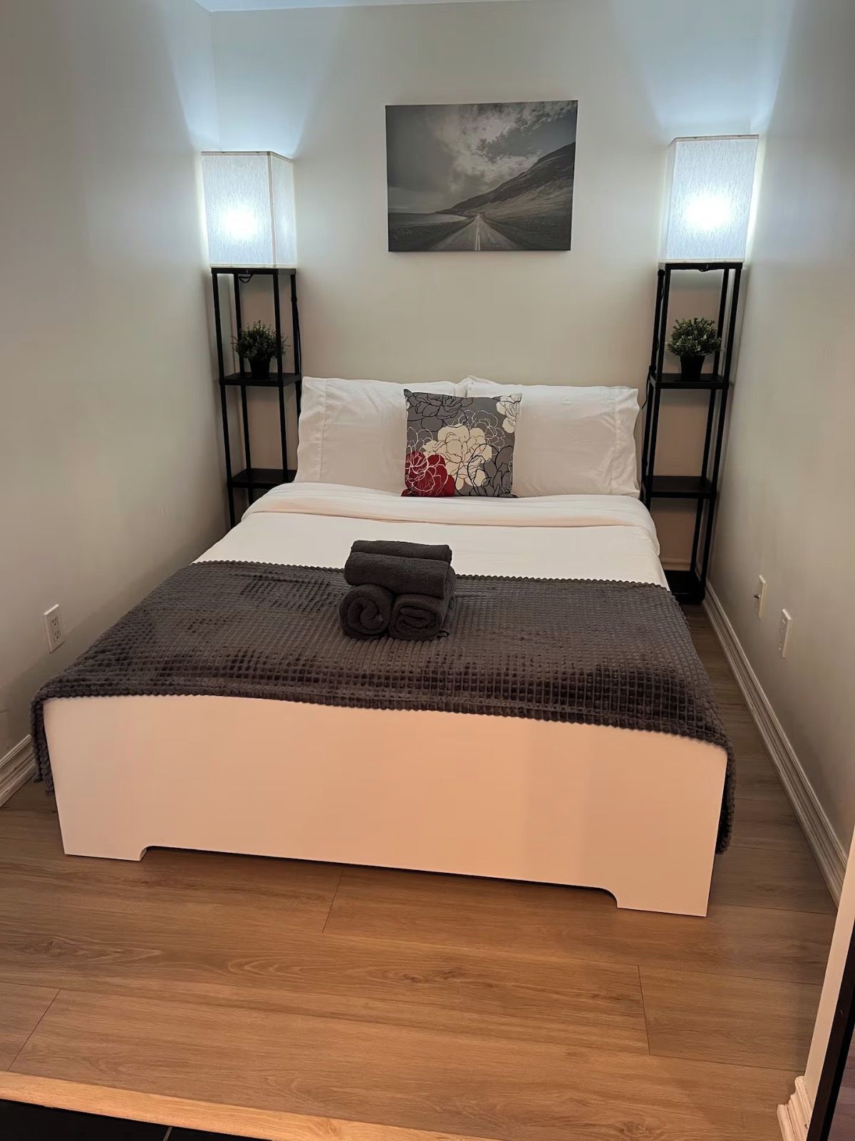 Un lit bien fait, recouvert d’une couette blanche et d’un plaid gris foncé, flanqué de deux tables de chevet noires avec des lampes, dans une pièce au plancher en bois et ornée d’un seul tableau accroché au mur.