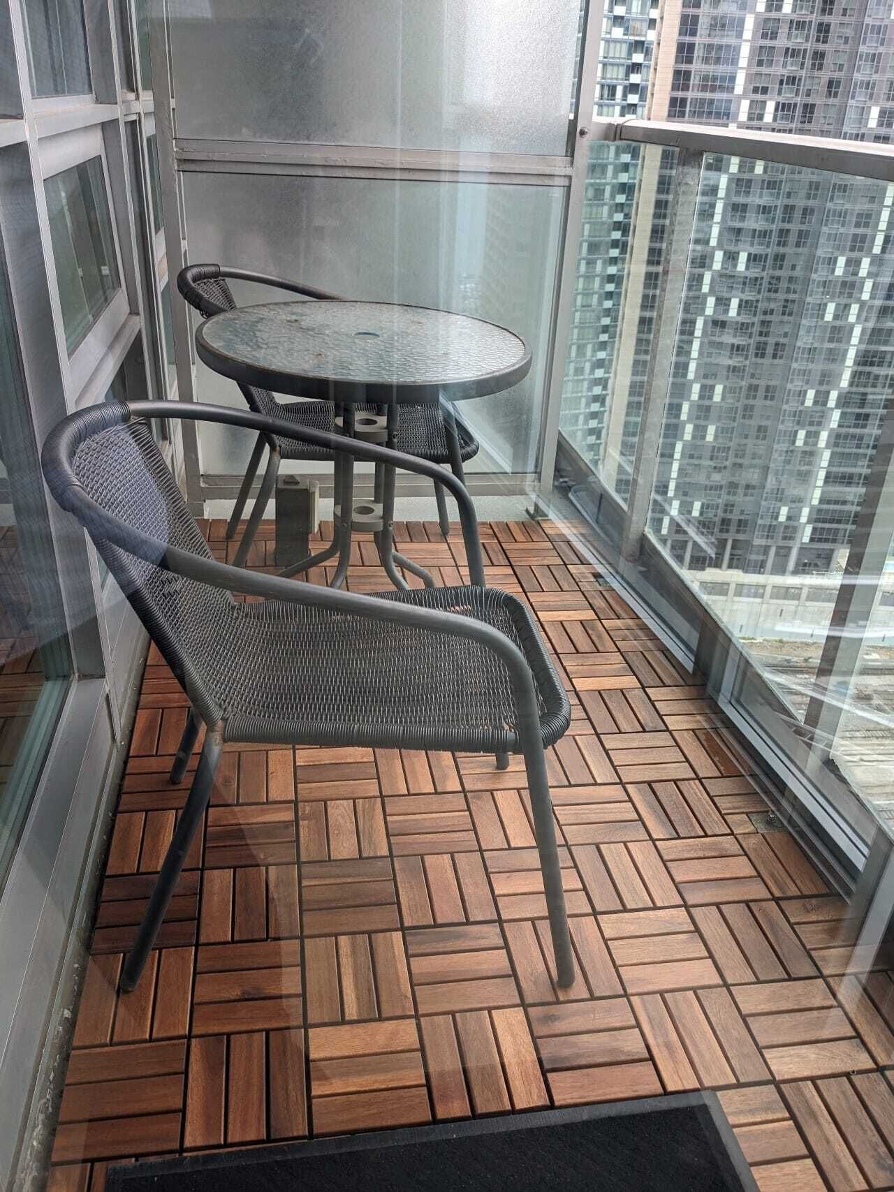 A balcony with a round table and two chairs, wooden flooring, and a view of a cityscape through large windows.