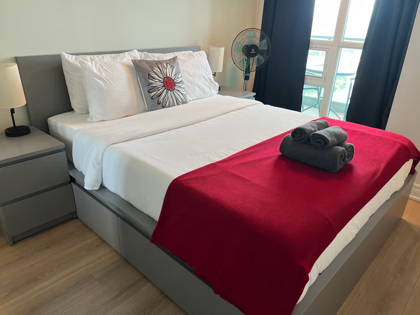 A neatly made bed with a red blanket, white pillows, and a single decorative pillow with a daisy design.