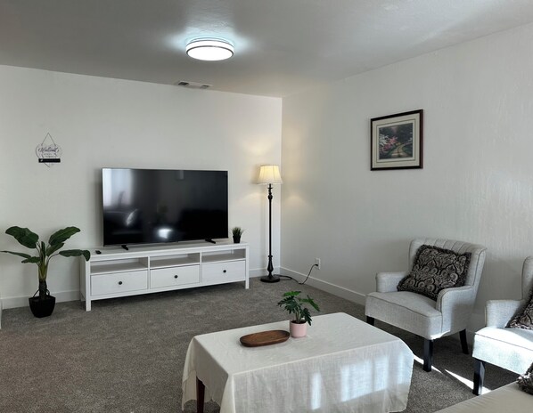 Ein Wohnzimmer mit Flachbildfernseher auf einer weißen Unterhaltungseinheit, einem Couchtisch mit Pflanze, einem grauen Sessel und einer Stehlampe.