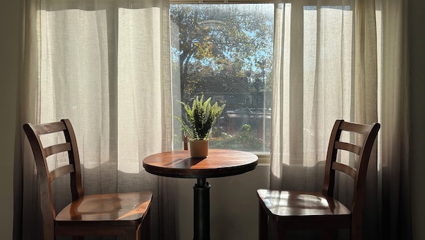 Una habitación con una mesa redonda de madera y dos sillas de madera, una planta en maceta sobre la mesa, y una ventana con cortinas traslúcidas.