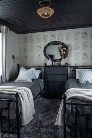 A bedroom with two beds, a dark ceiling, a patterned wallpaper, a mirror, and a wall-mounted lamp.