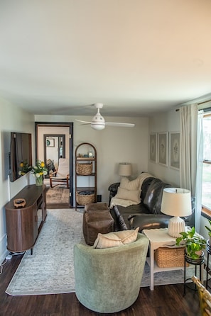 A living room with a sofa, armchair, and a small table with a lamp. There is a mirror on the wall, a ceiling fan, and a door leading to another room.