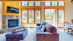 A spacious living room with a stone fireplace, a flat-screen TV, a large window, and a dining area. - Gateway to Crater Lake! Resort Retreat w/ Pool (Klamath Falls)