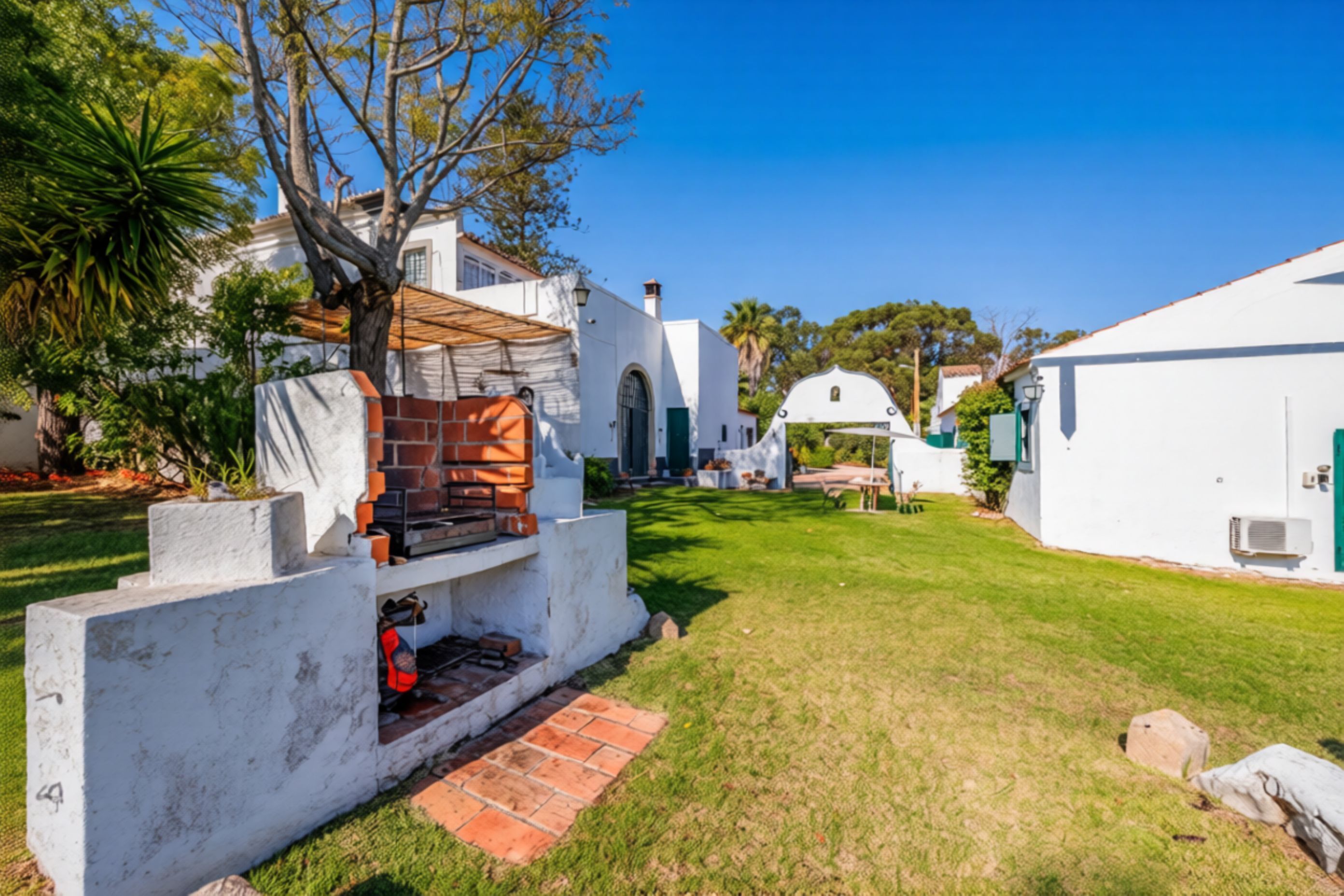 A white building with a brick barbecue area, a small garden, and a clear blue sky.