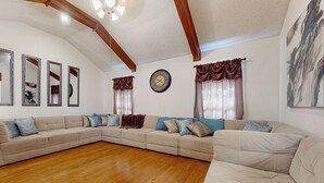 Un ampio soggiorno con un grande divano beige a forma di L, pavimento in legno e soffitto con travi a vista.