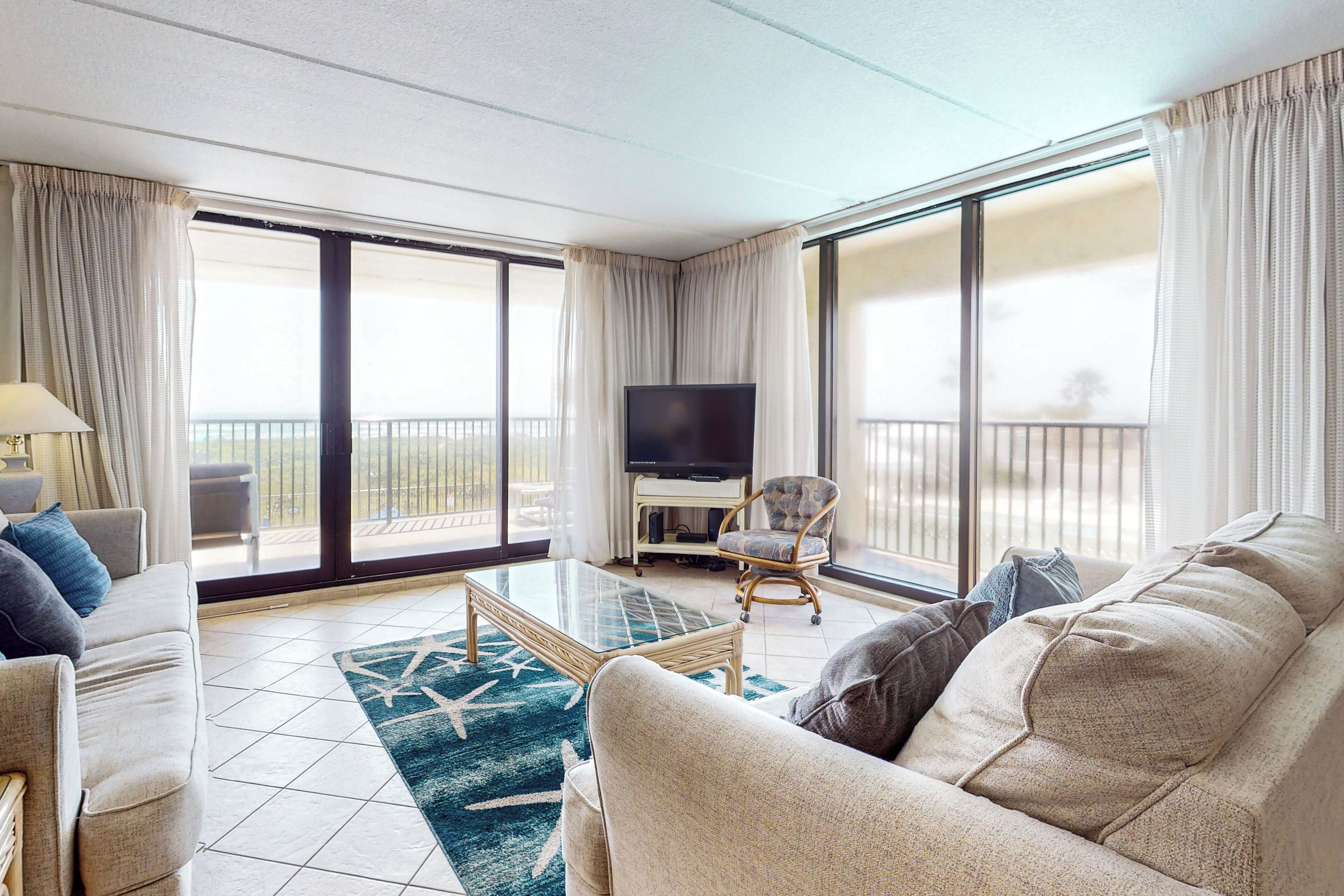 Ein geräumiges Wohnzimmer mit einer Couch, einem Sessel und einem Glastisch. Ein Flachbildfernseher ist an der Wand befestigt, und es gibt einen Balkon mit Blick.