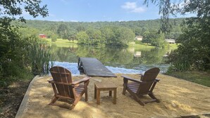 Une terrasse en bois avec deux chaises et une table, d’où l’on a vue sur un lac, le tout entouré d’une végétation luxuriante et de maisons situées au loin.